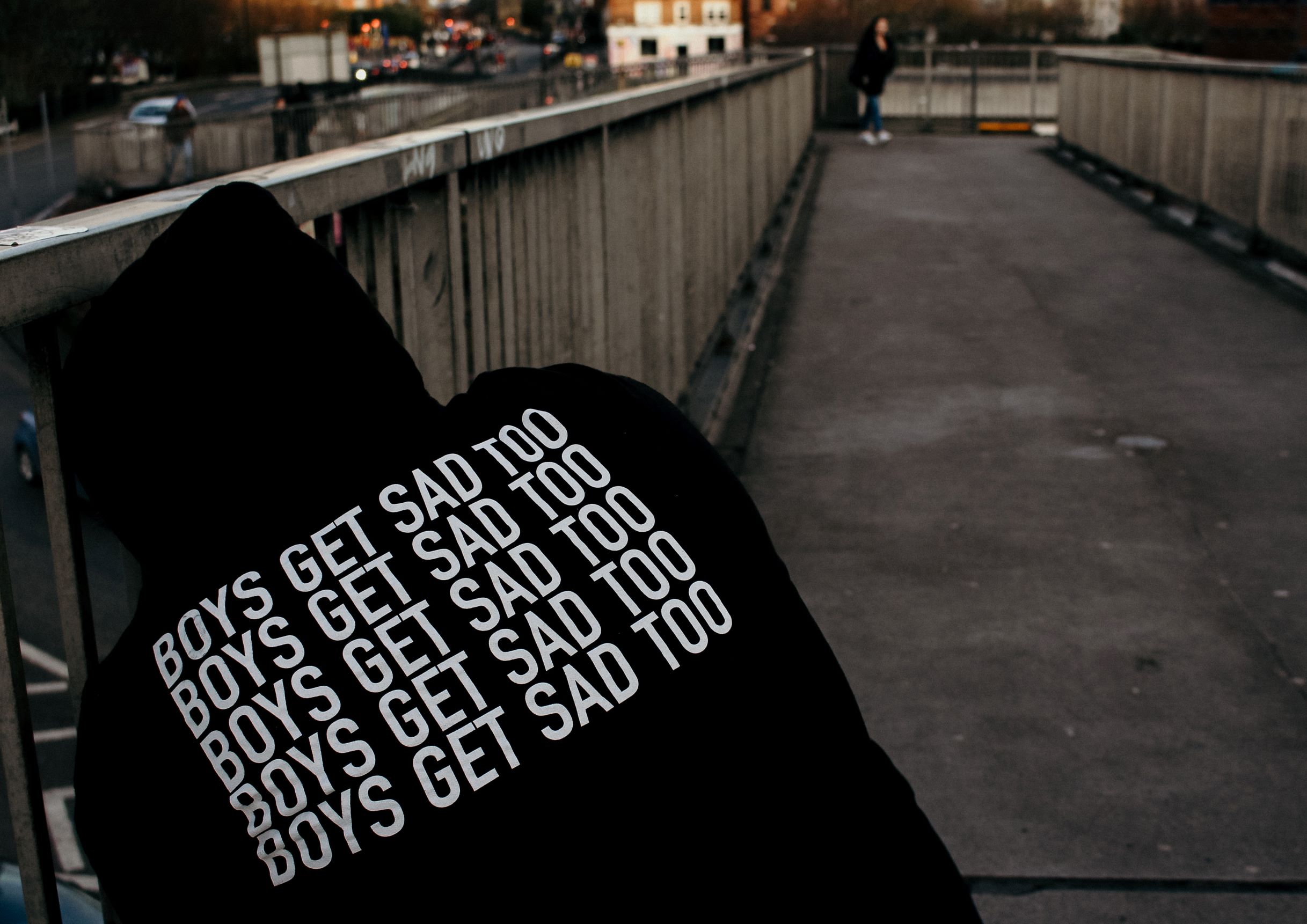 Back of a person wearing a black sweatshirt that says "Boys Get Sad Too." Colors are dark to represent depression in men.